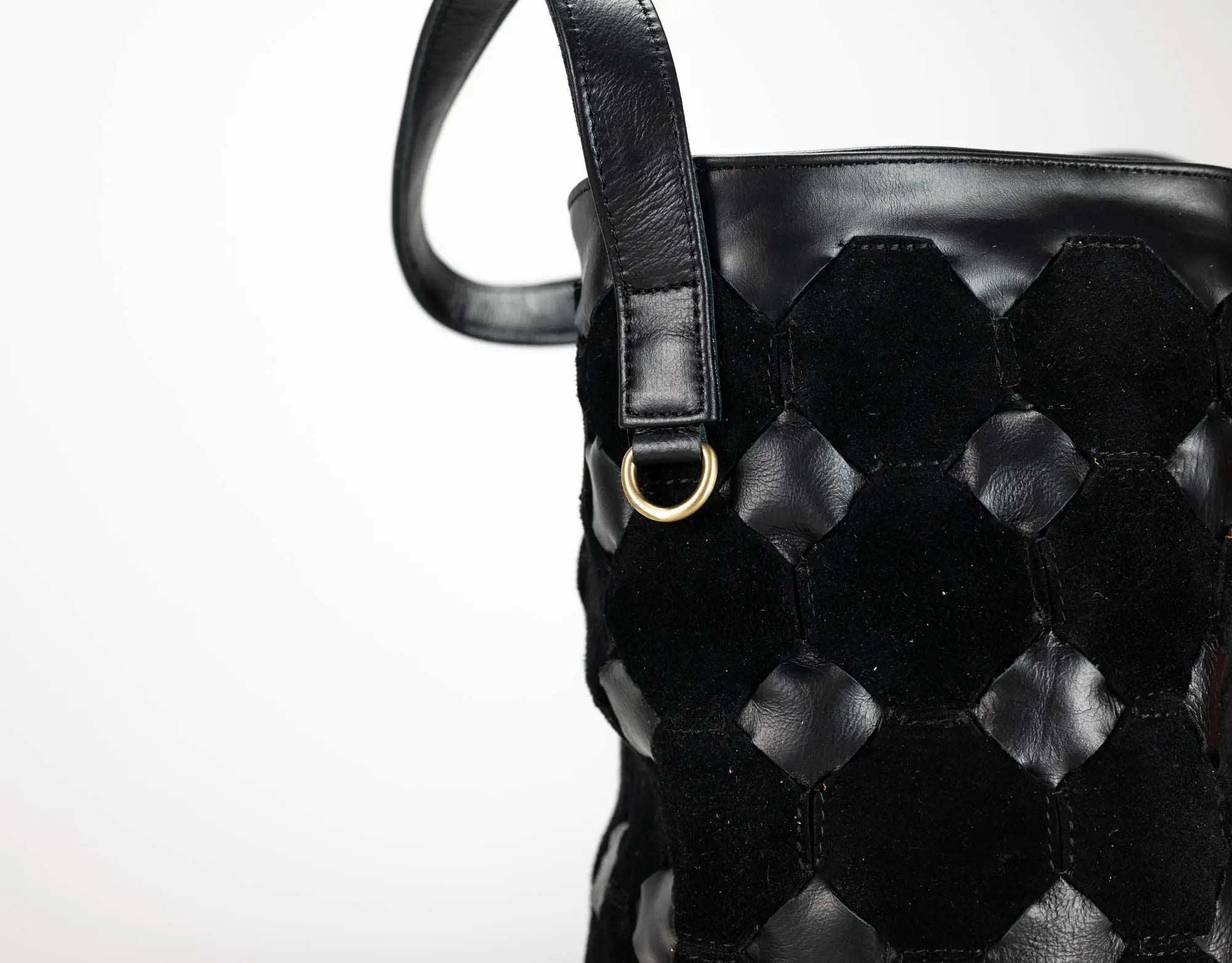 Close-up of a black woven leather handbag with a hexagonal pattern, a black leather strap, and a small gold-tone metal ring on a white background.