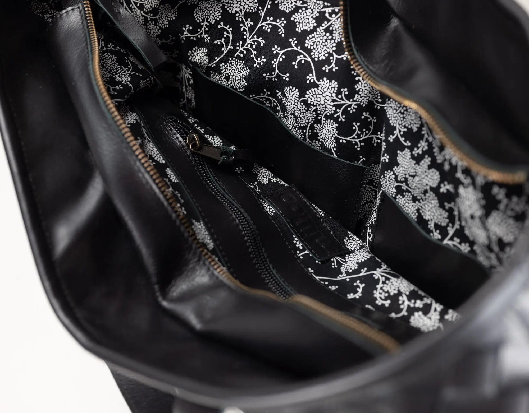 Close-up of the interior of a black bag, showing a black fabric lining with a white floral damask pattern and gold-toned zipper trim.