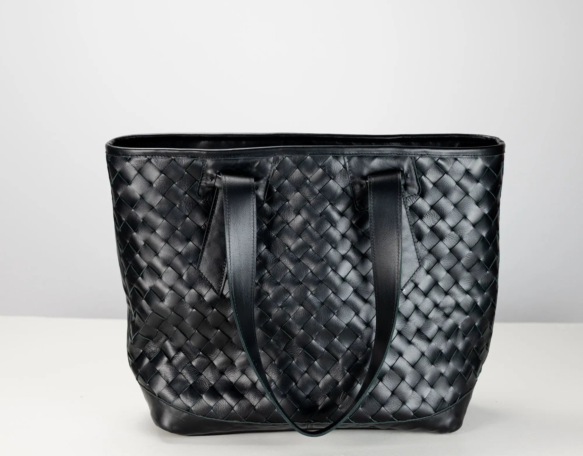 A black woven leather tote bag with two shoulder straps, photographed against a plain light gray background.