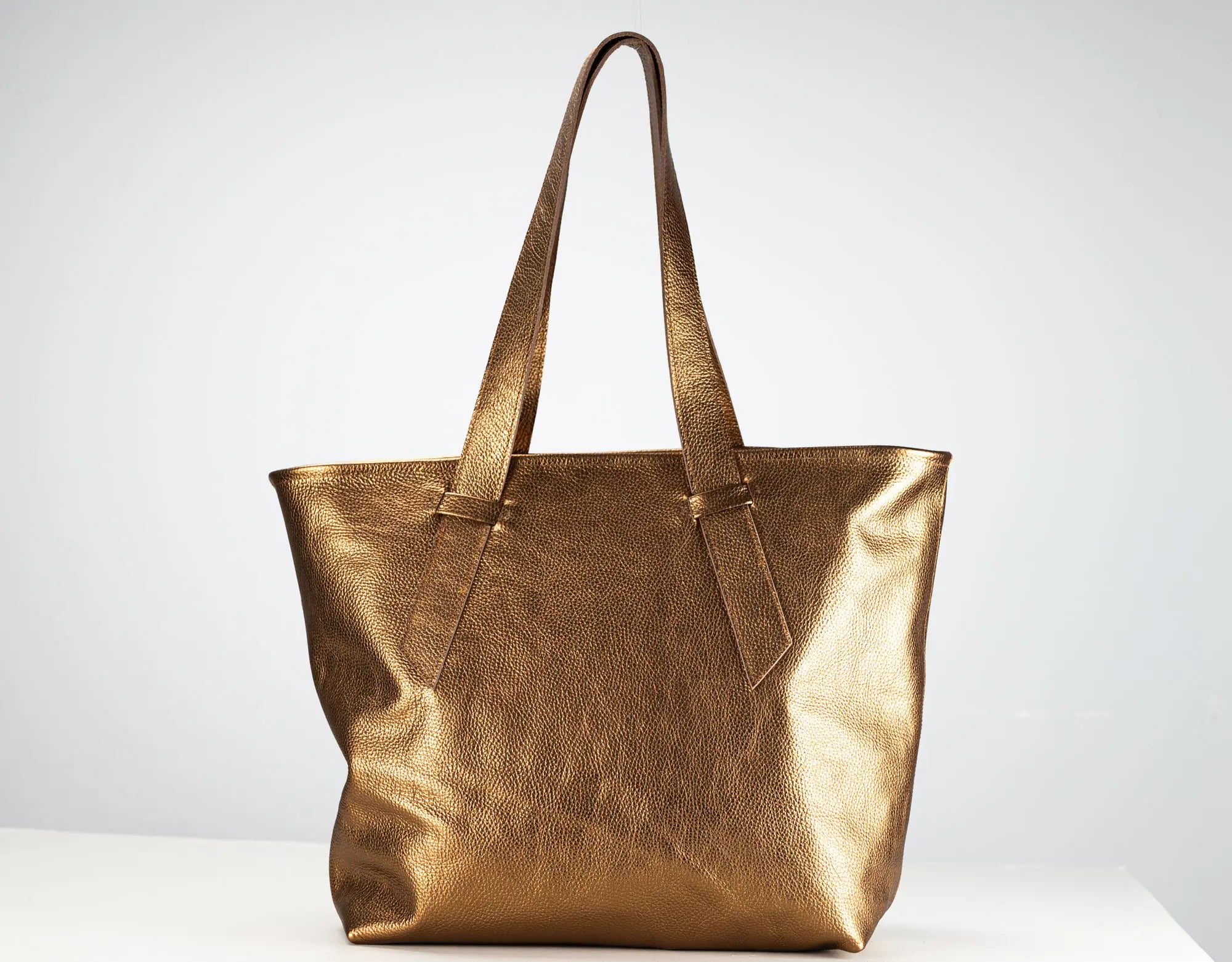 A metallic gold leather tote bag with dual shoulder straps, standing upright on a white surface against a light gray background.