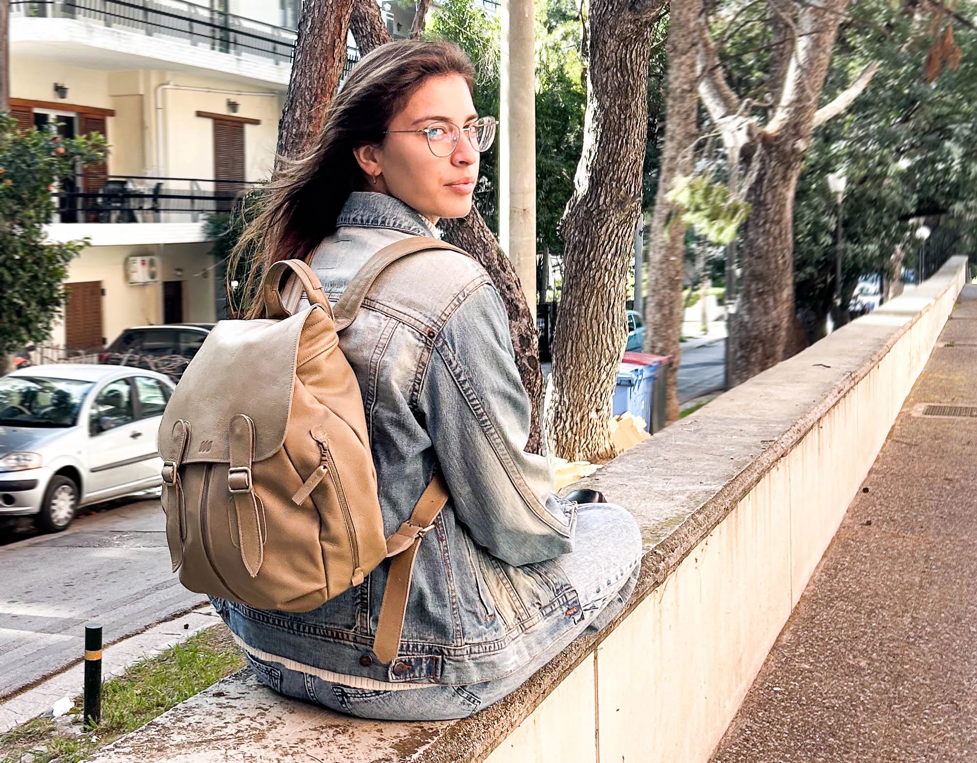 Woman with a beige backpack sitting on a ledge by a street.