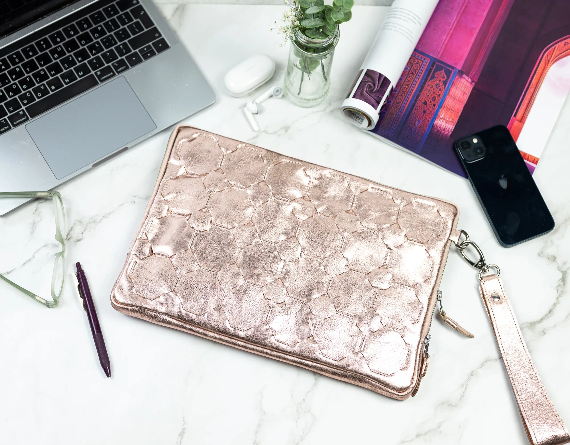 Rose gold clutch on a marble surface with laptop, phone, and glasses.