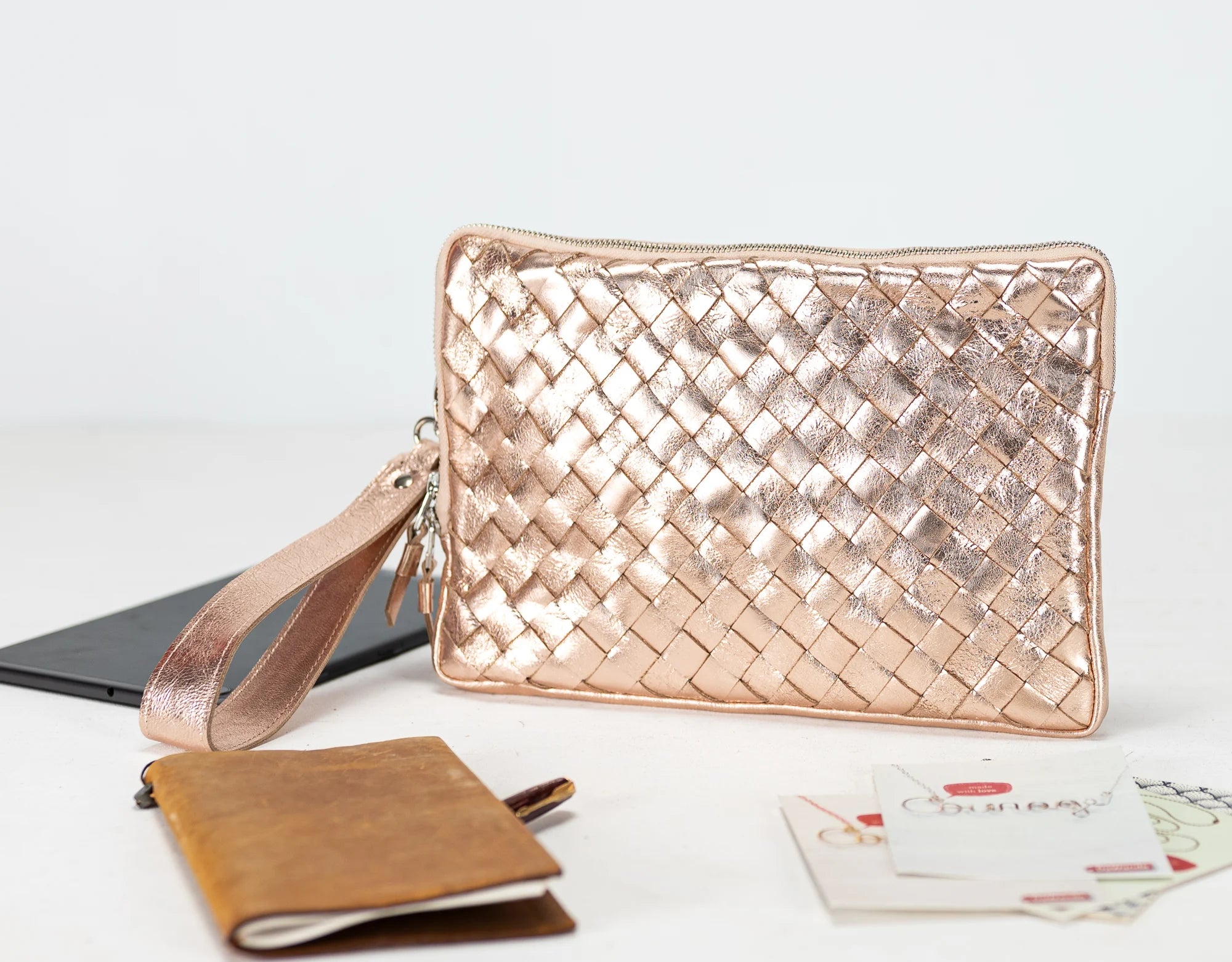Rose gold woven clutch bag on a light surface with a phone and wallet.