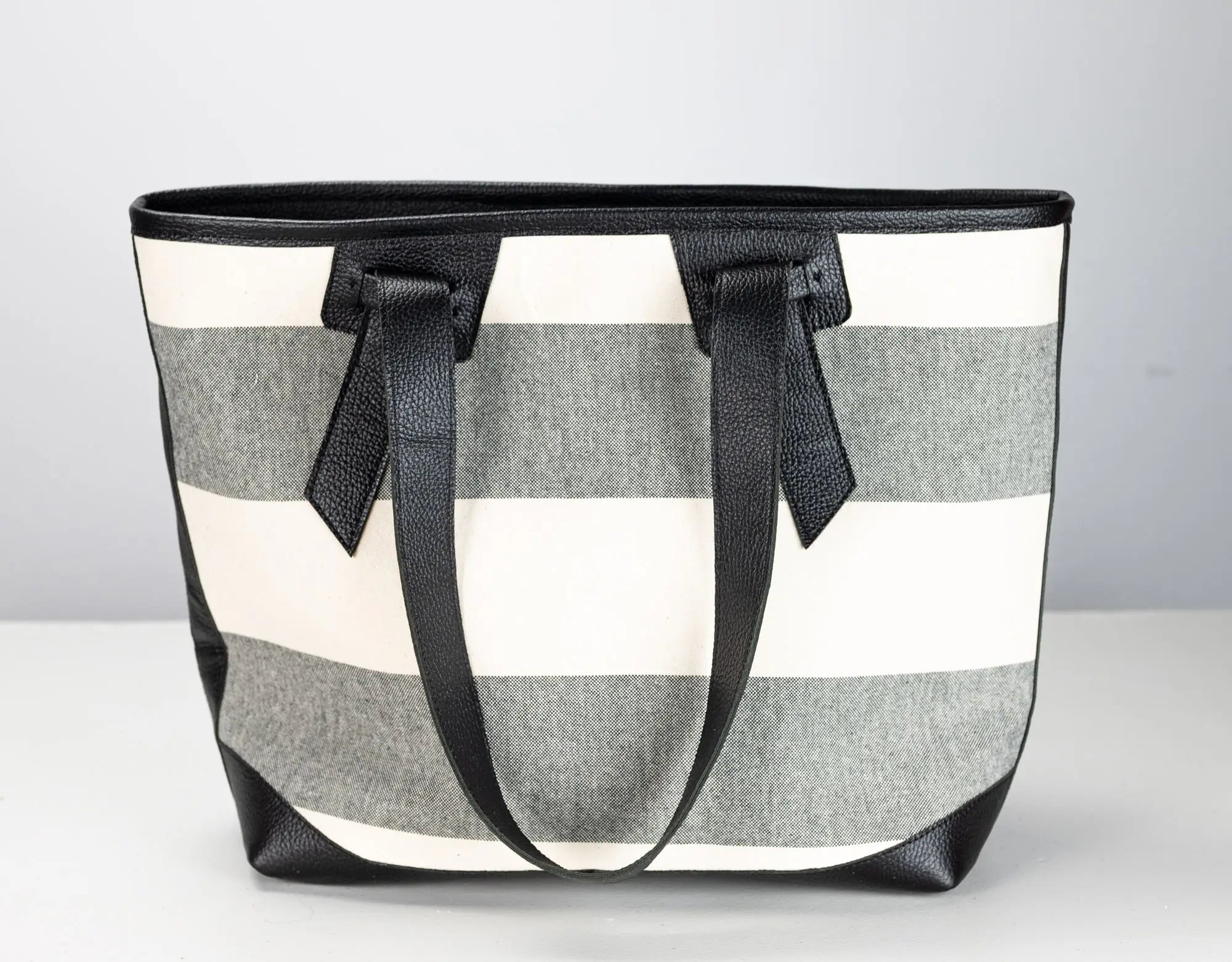 A striped tote bag with horizontal gray and off-white stripes, featuring black leather handles and trim, set against a neutral light gray background.