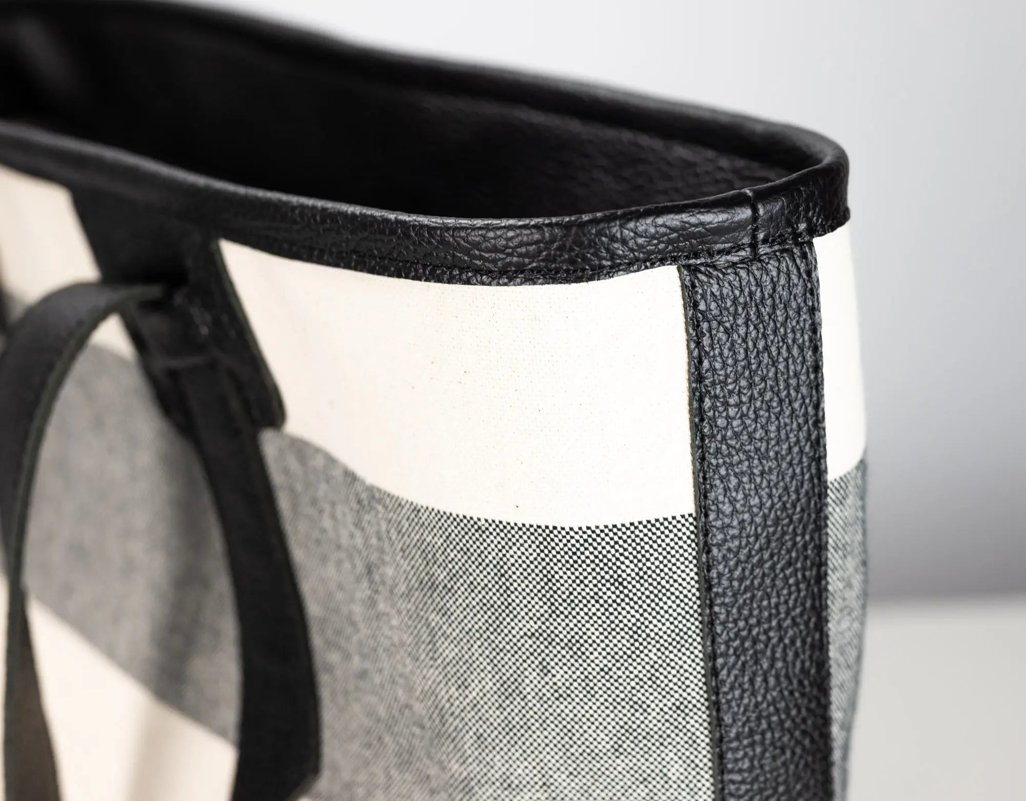 Close-up of a tote bag corner showing black leather trim, a black handle, and a black-and-white check patterned fabric body.