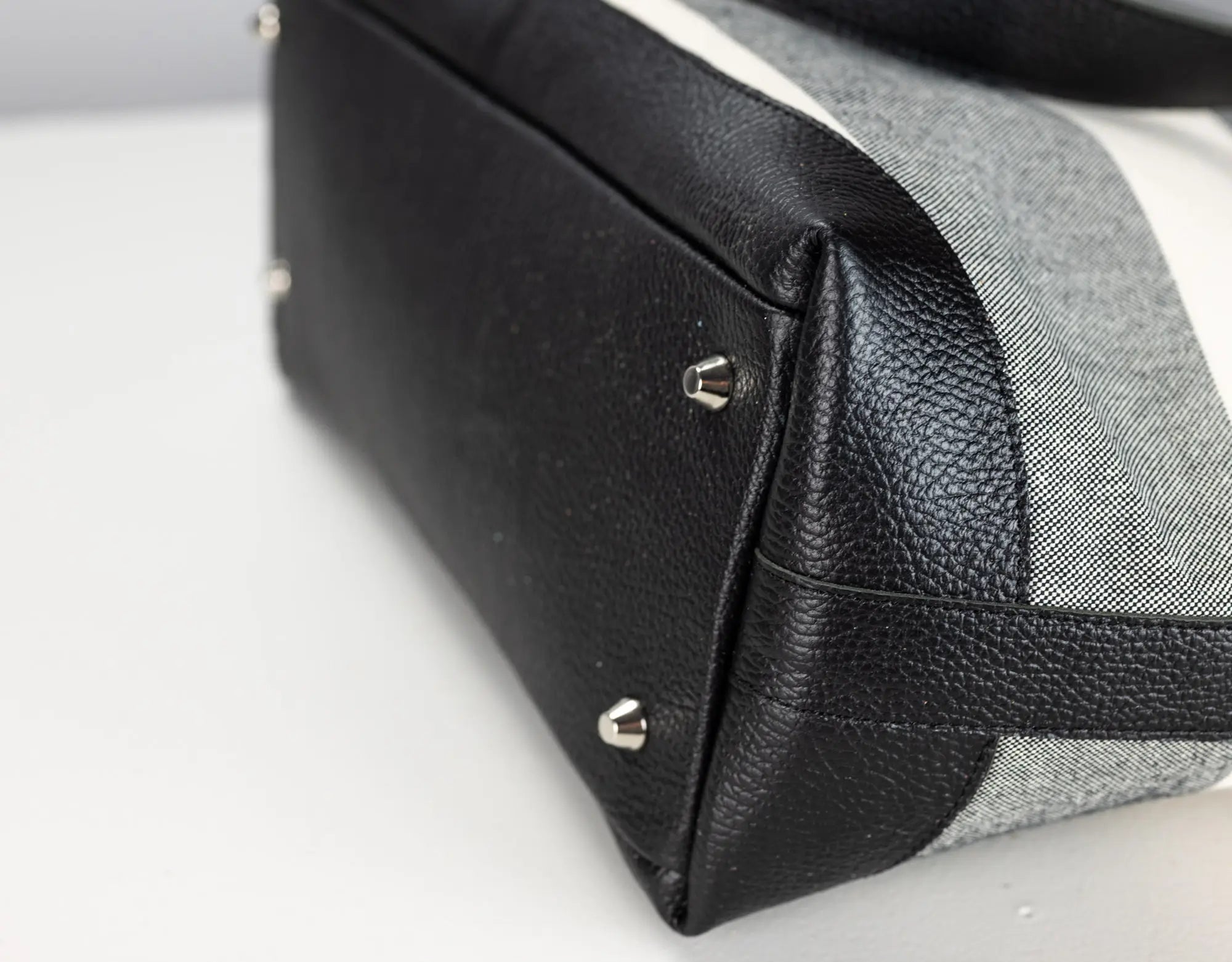 Close-up of a handbag corner showing black pebbled leather trim and silver-tone metal studs against a light gray and white woven fabric body.