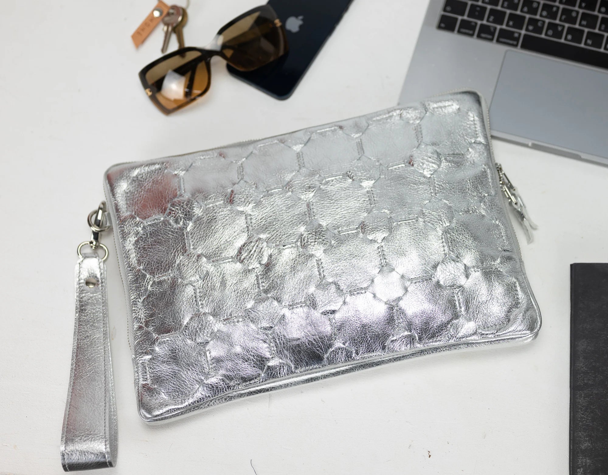 Silver clutch bag on a white surface with sunglasses, phone, and laptop in the background