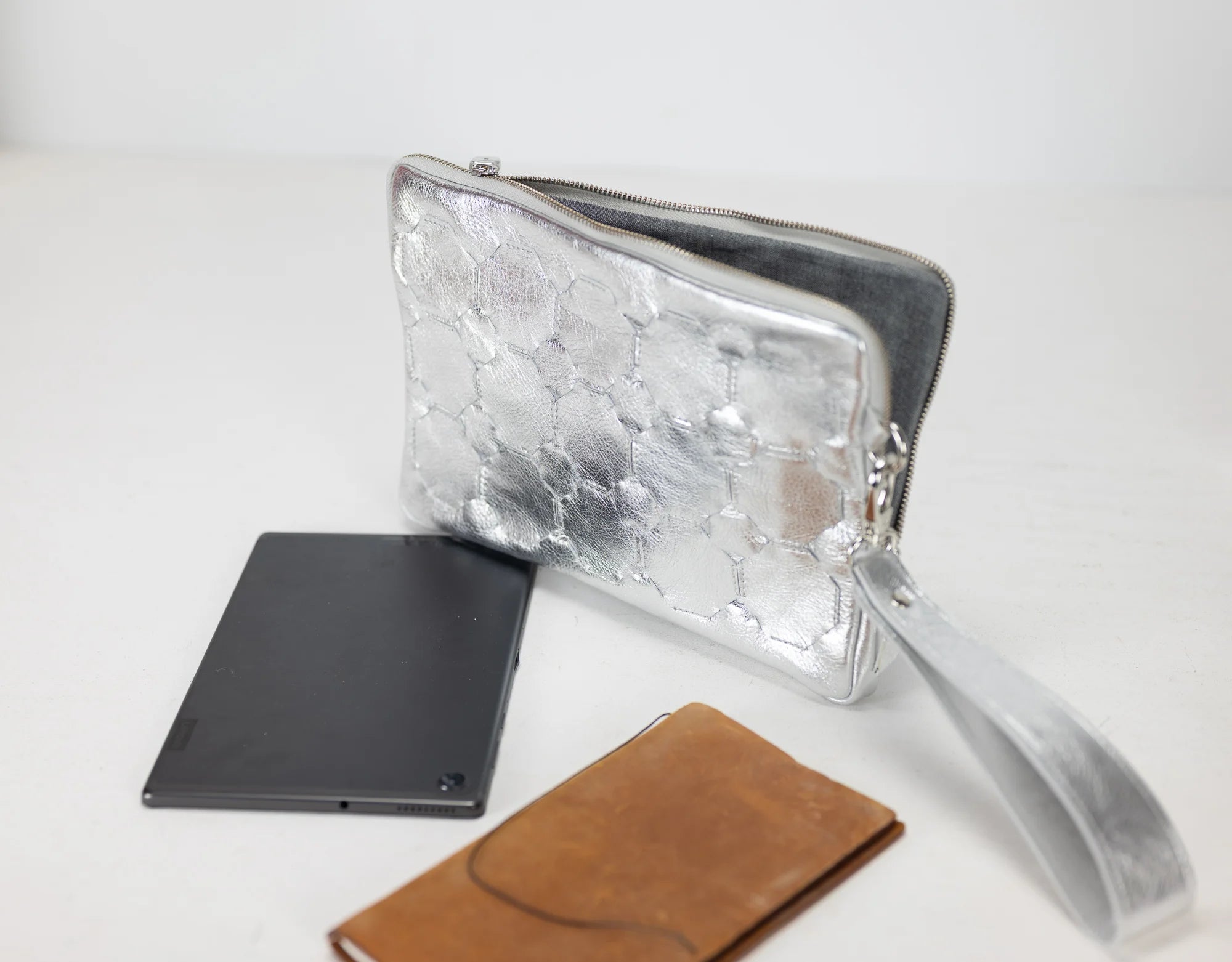 Silver clutch bag with a black tablet and brown wallet on a white background