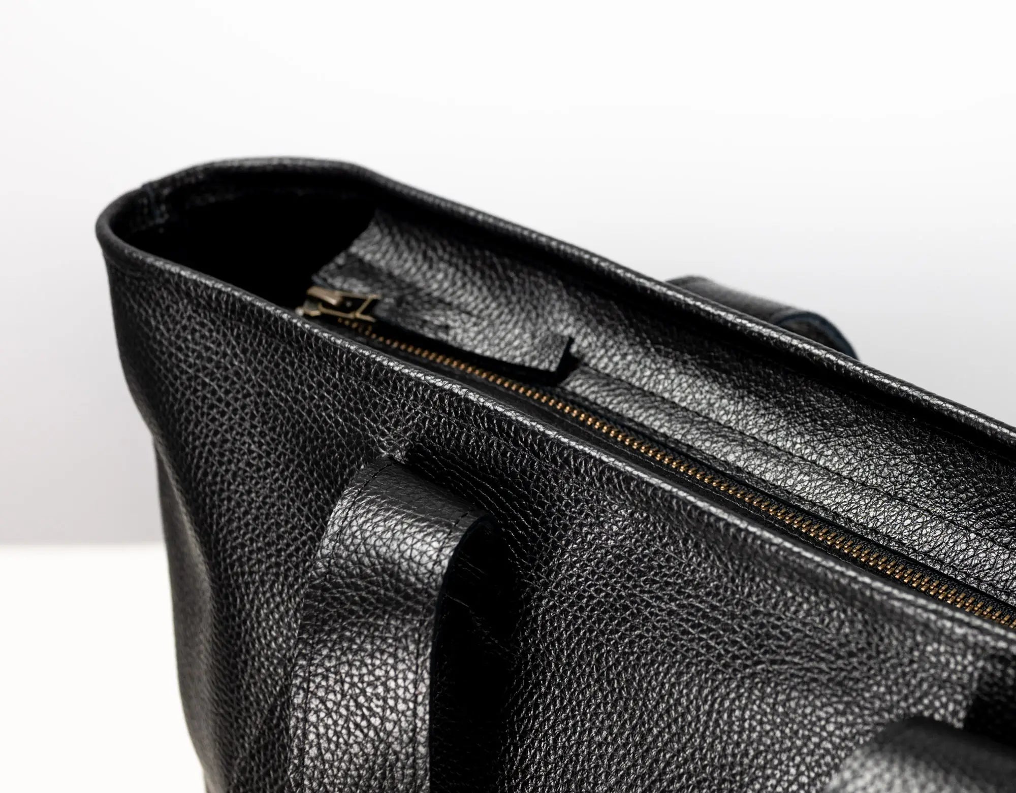 Close-up of a black pebbled leather bag showing the interior opening with a gold-toned zipper and a handle attachment, photographed against a clean white background.