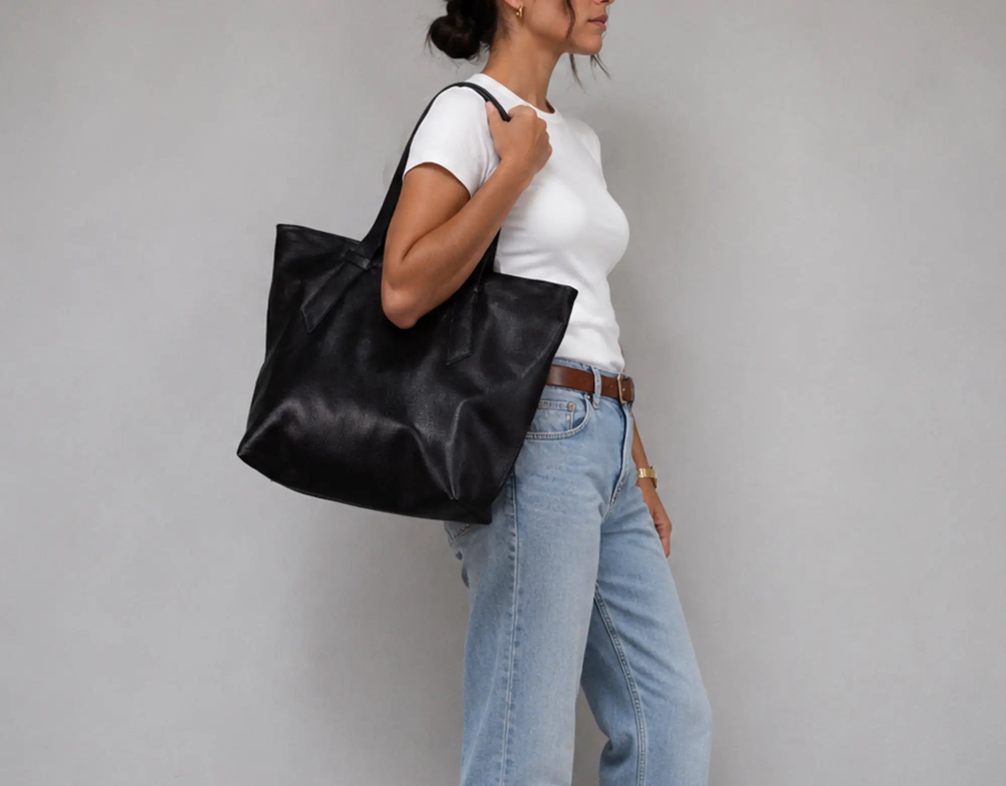 A woman wearing a white t-shirt, light blue jeans, and a brown belt carries a large black leather tote bag over her shoulder against a plain gray studio background.