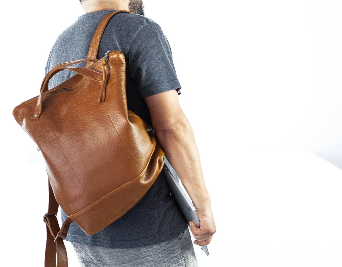 Person wearing a brown leather backpack and holding a laptop on a white background