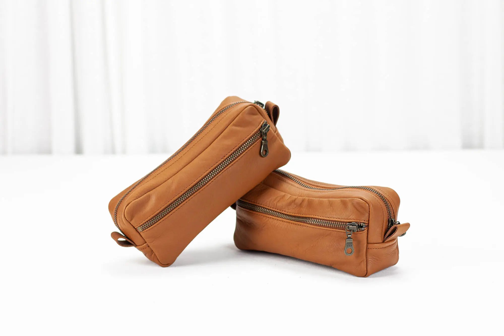 Two brown leather toiletry bags with zippers on a white background