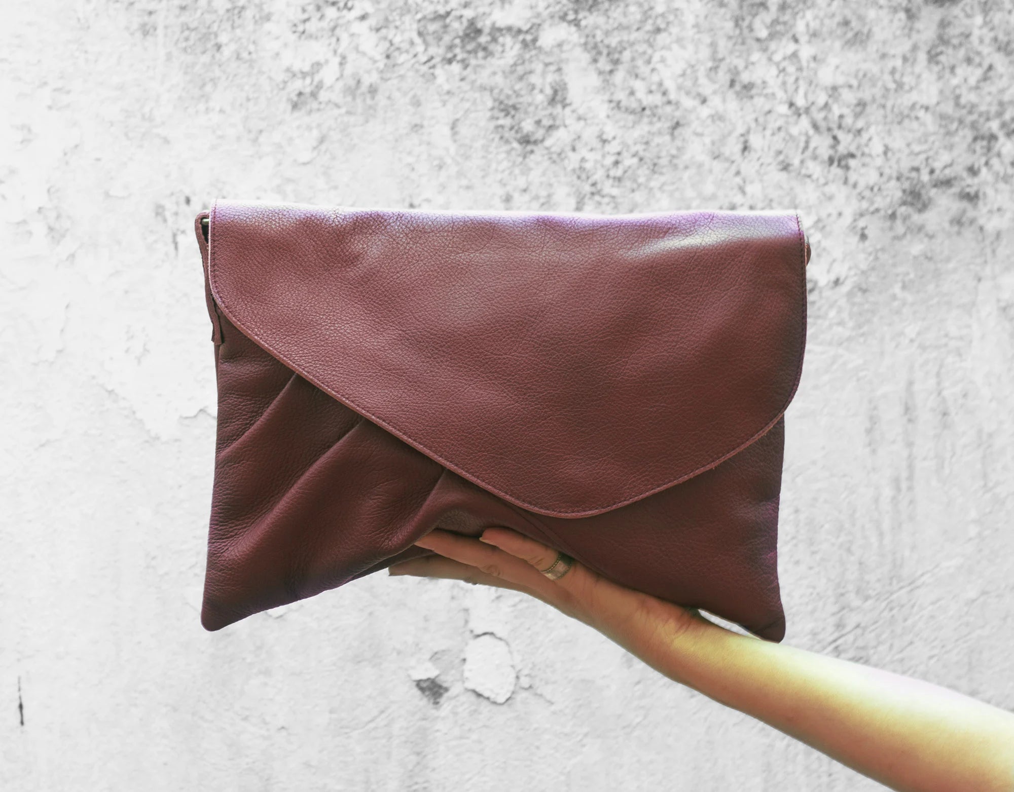 Burgundy leather clutch held by a hand against a textured white background