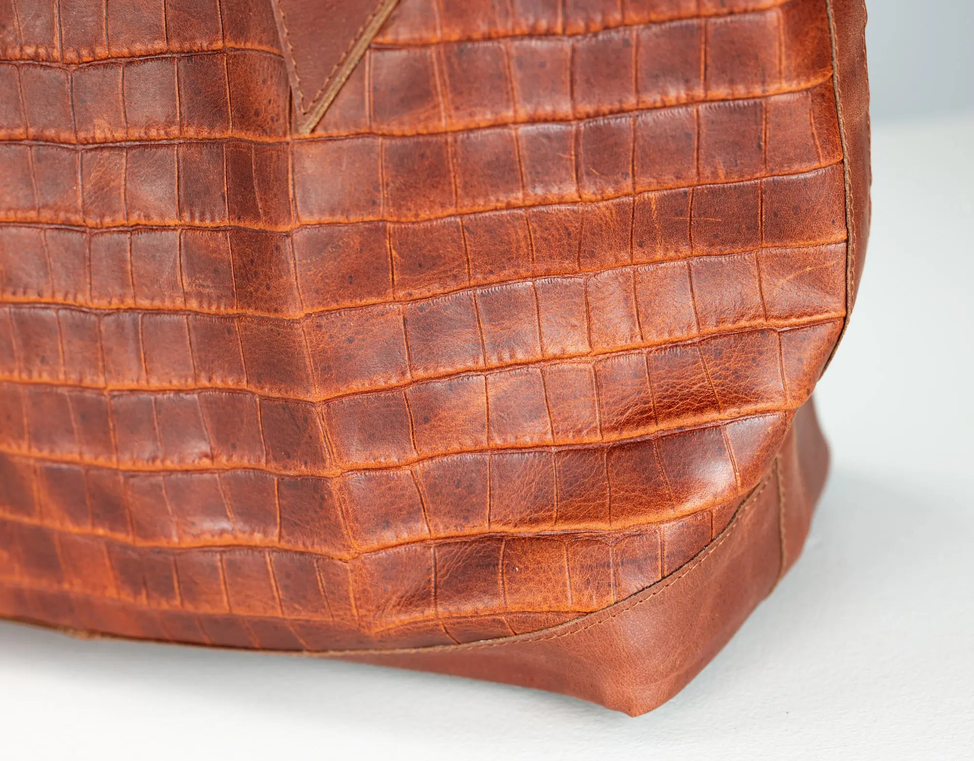 Close-up of a brown crocodile-embossed leather bag corner showing the textured pattern and stitched edge on a light gray background.