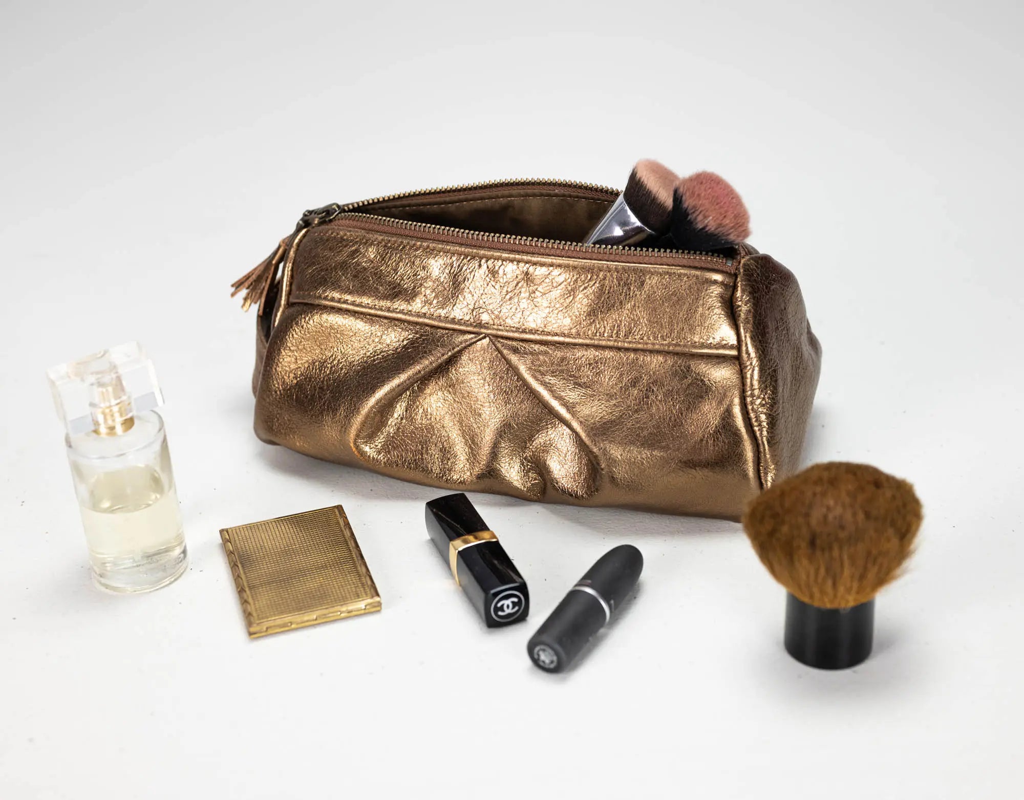 A metallic gold makeup bag with a zippered top, shown open with makeup brushes inside, alongside cosmetic items including a perfume bottle, compact powder, lipsticks, and a large makeup brush on a white background.