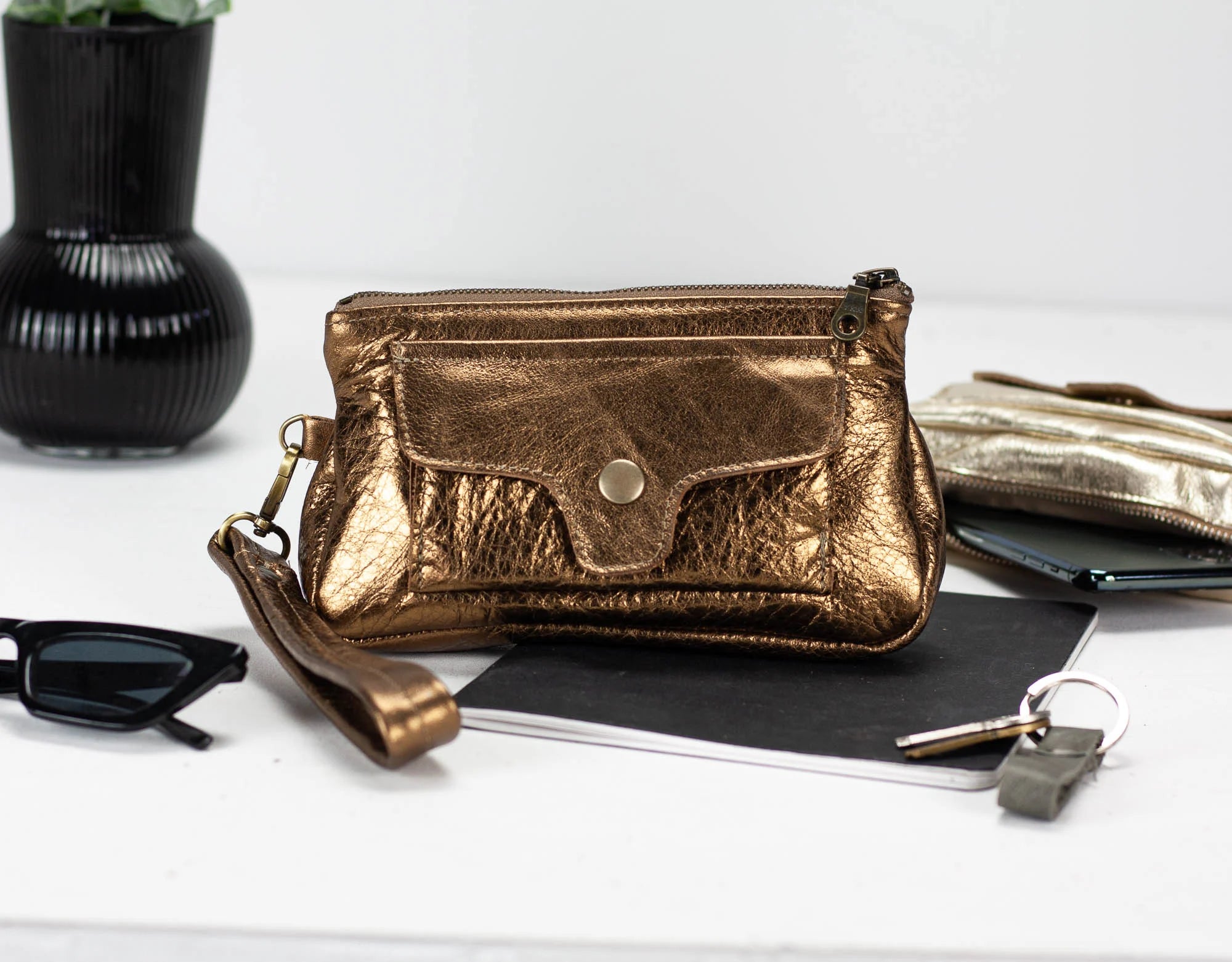 Brass leather clutch bag on a white surface with sunglasses and a notebook.