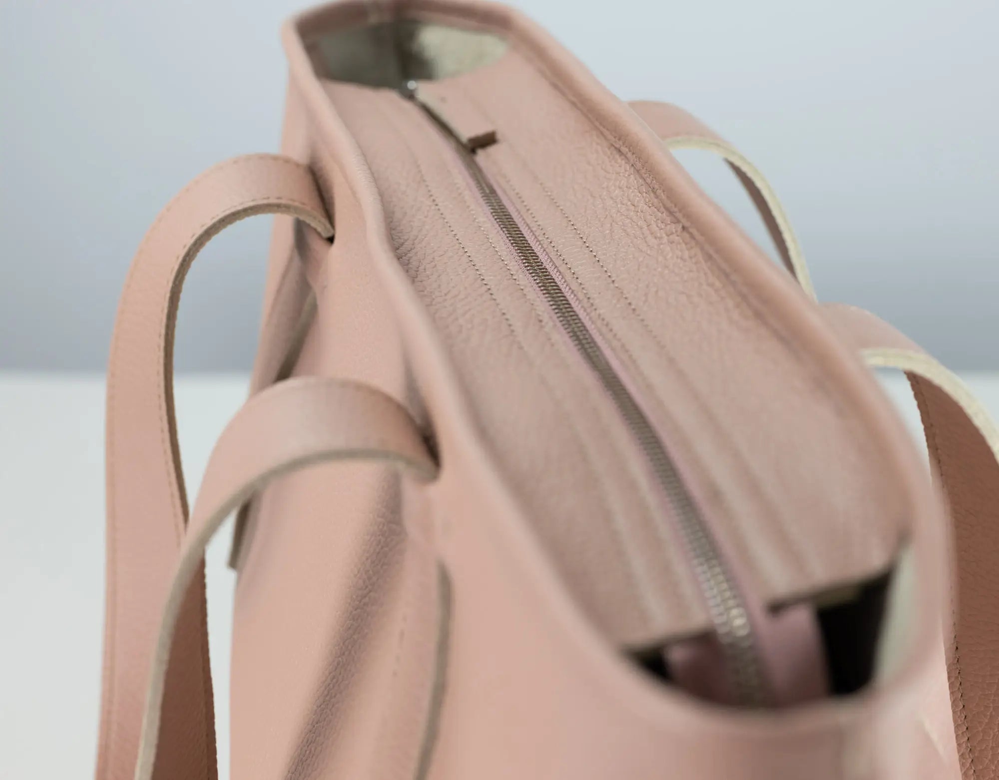 Close-up of a blush pink pebbled leather tote bag showing the interior zippered compartment and side handles.