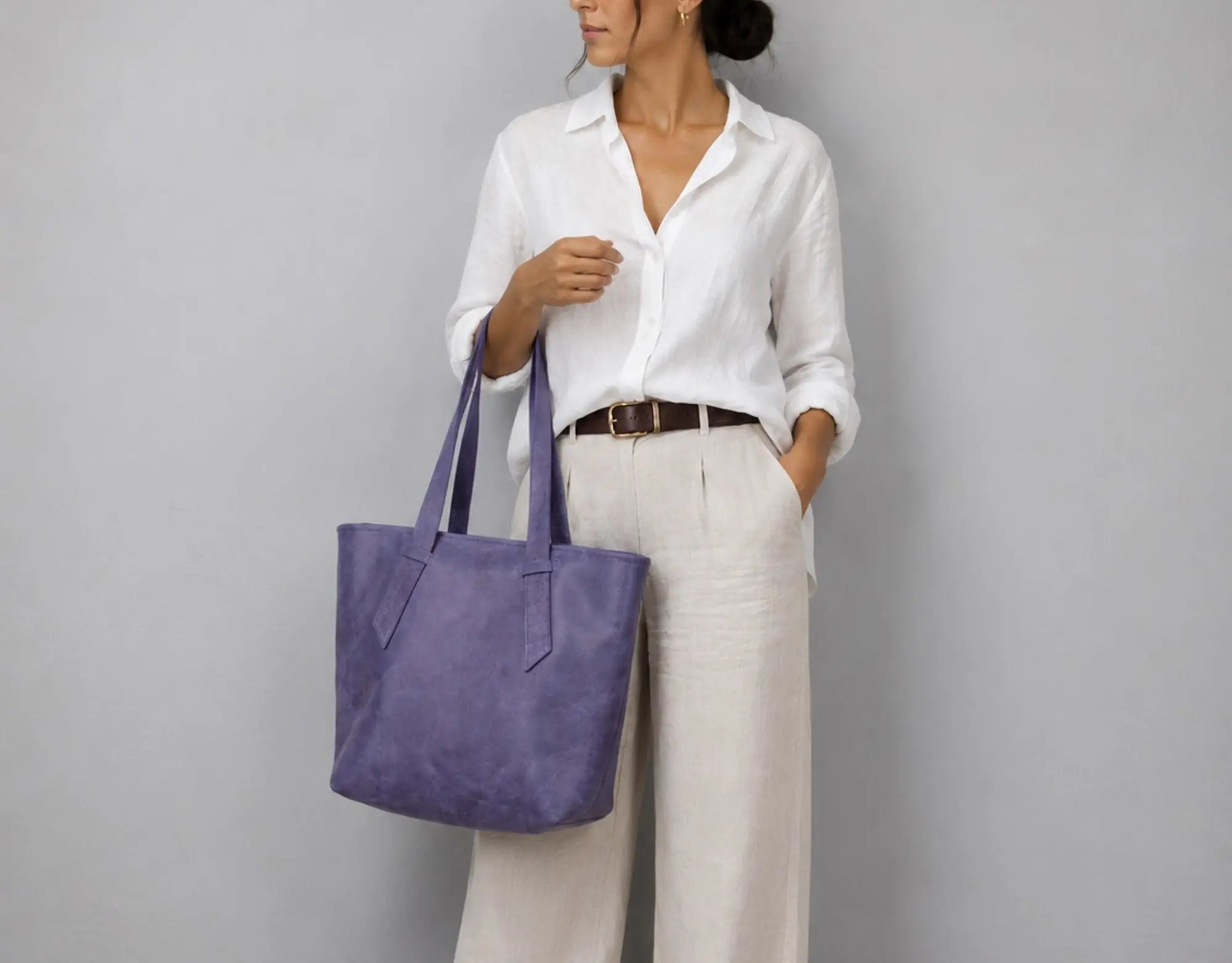 A woman wearing a white button-down shirt and cream trousers carries a large lavender-purple leather tote bag against a plain gray studio background.