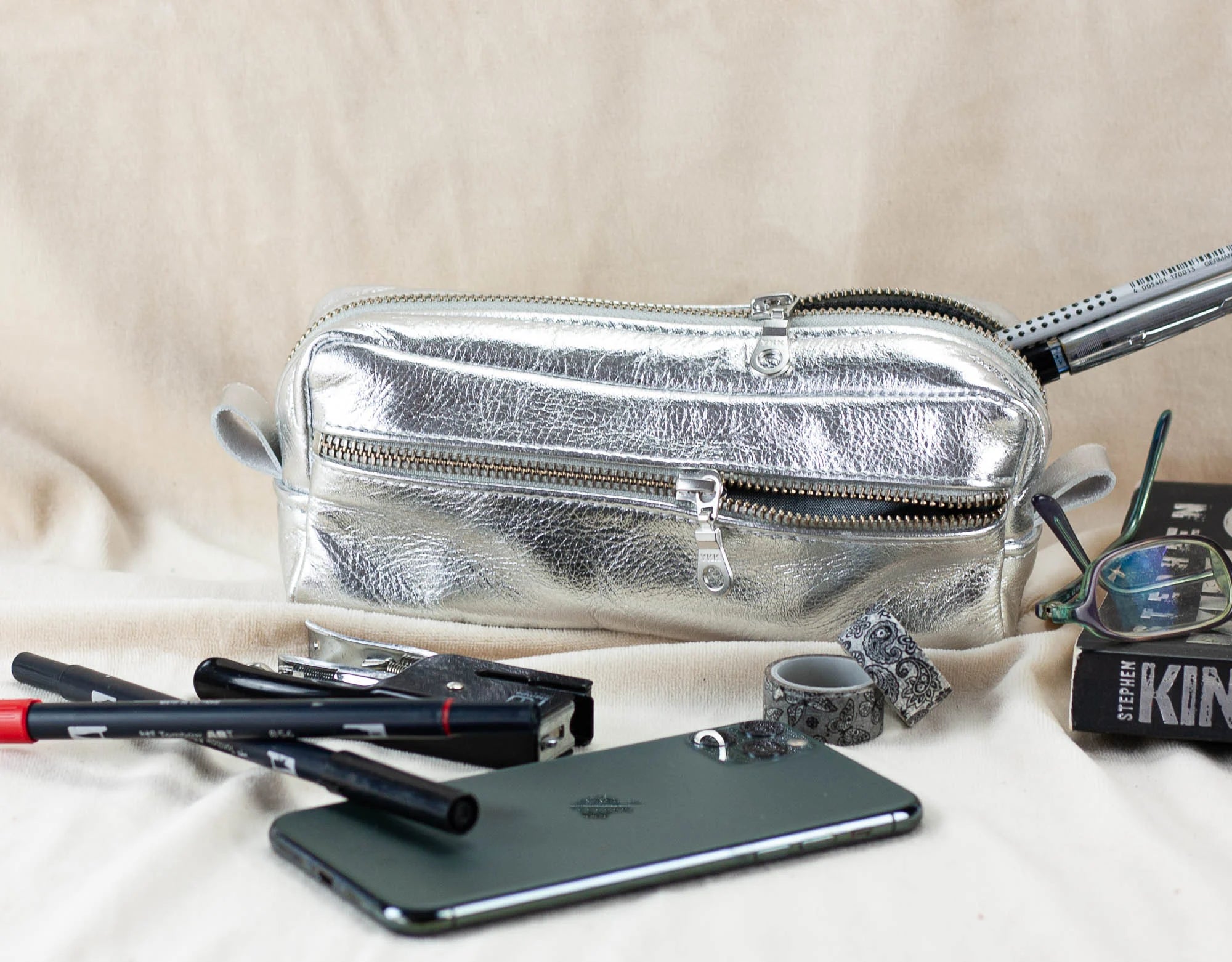 Silver fanny pack with makeup items and a phone on a beige surface