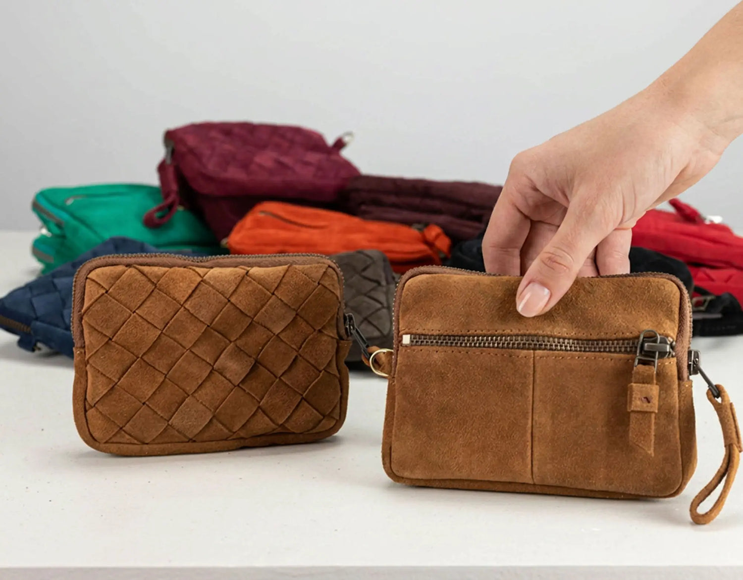 Two small brown crossbody wallets are shown on a white surface, with one hand holding the right wallet. A stack of other colorful wallets sits blurred in the background.