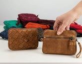 Two small brown crossbody wallets are shown on a white surface, with one hand holding the right wallet. A stack of other colorful wallets sits blurred in the background.