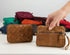 Two small brown crossbody wallets are shown on a white surface, with one hand holding the right wallet. A stack of other colorful wallets sits blurred in the background.