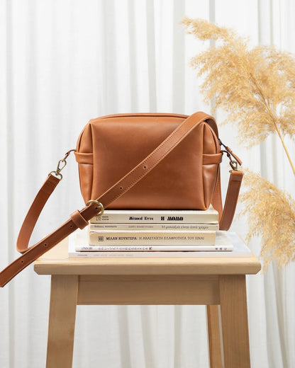 Brown leather bag on a wooden stool with books underneath, against a white curtain background.