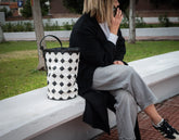 Person sitting on a bench with a patterned bag next to them, outdoors.