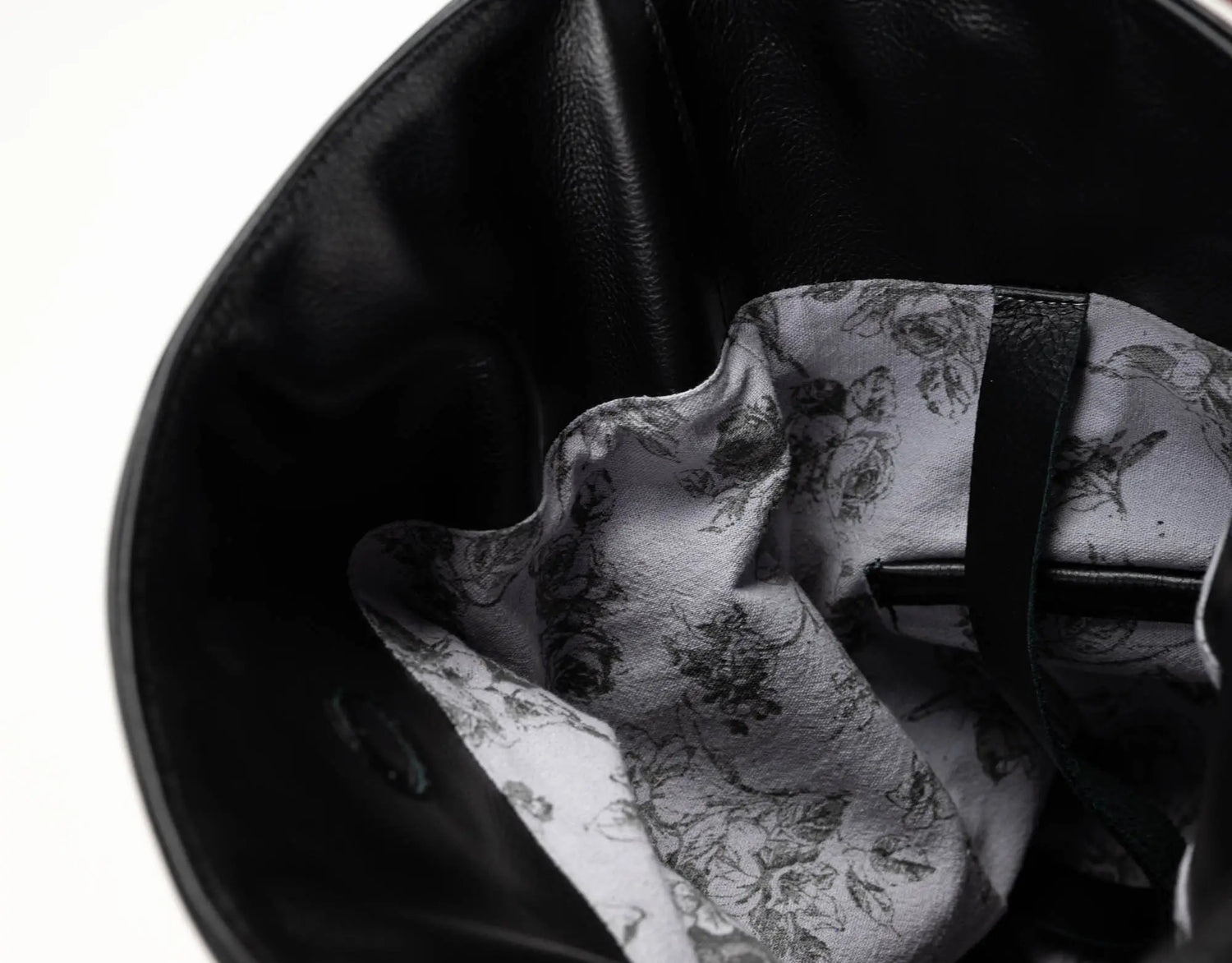 Close-up of the interior of a black leather bag, showing a white lining with a black floral and bird pattern and a black strap.