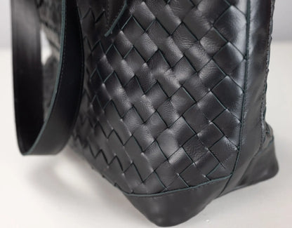 Close-up of a black woven leather handbag showing the detailed basketweave texture and black strap on a light gray background.