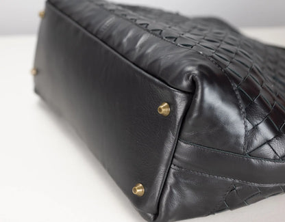 Close-up of a black leather handbag showing the bottom corner with gold-tone studs and a textured woven leather pattern on the side, set against a plain white background.