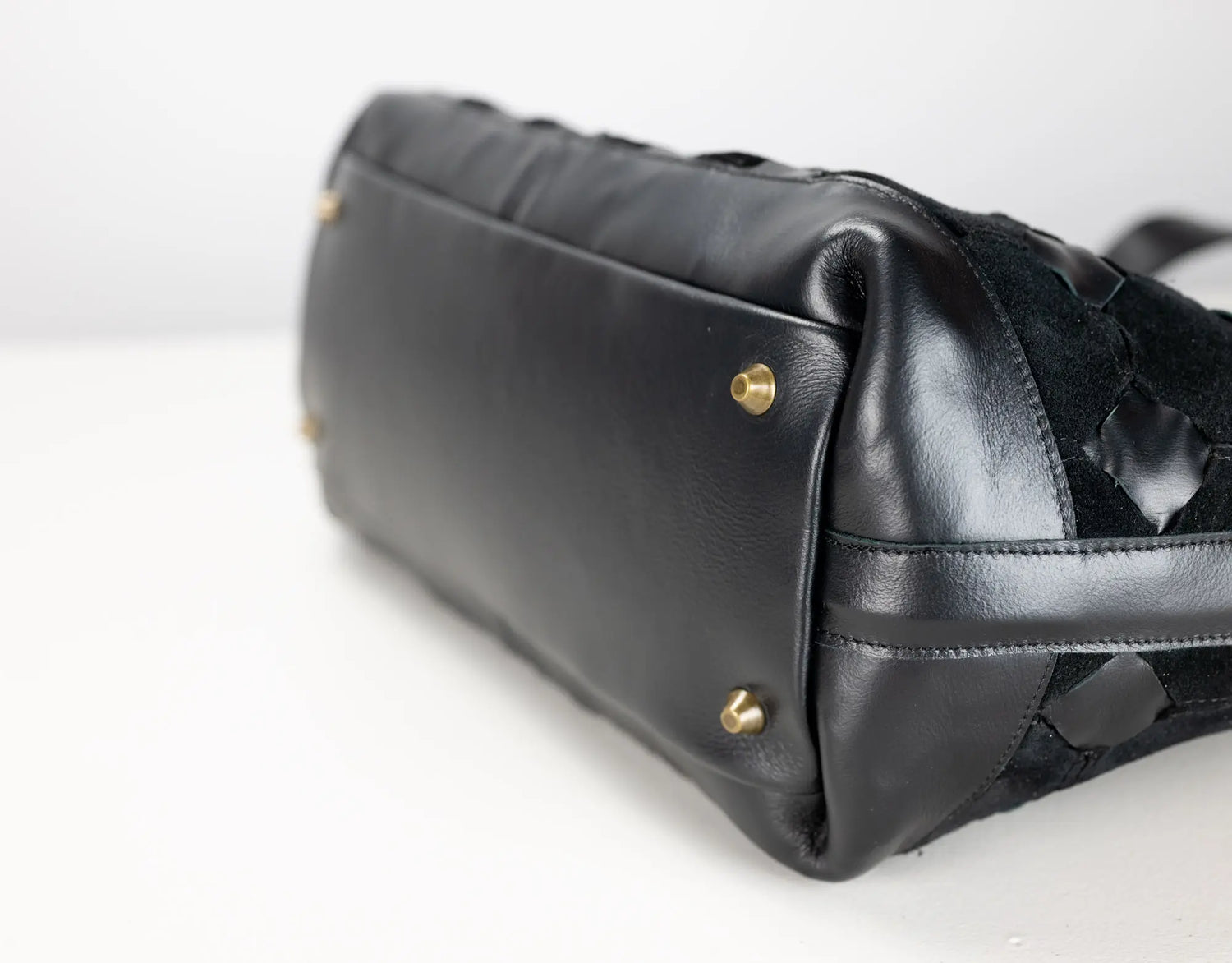 Close-up of a black leather handbag showing the bottom corner with gold-tone studs and a textured woven black strap detail on a white background.
