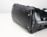 Close-up of a black leather handbag showing the bottom corner with gold-tone studs and a textured woven black strap detail on a white background.