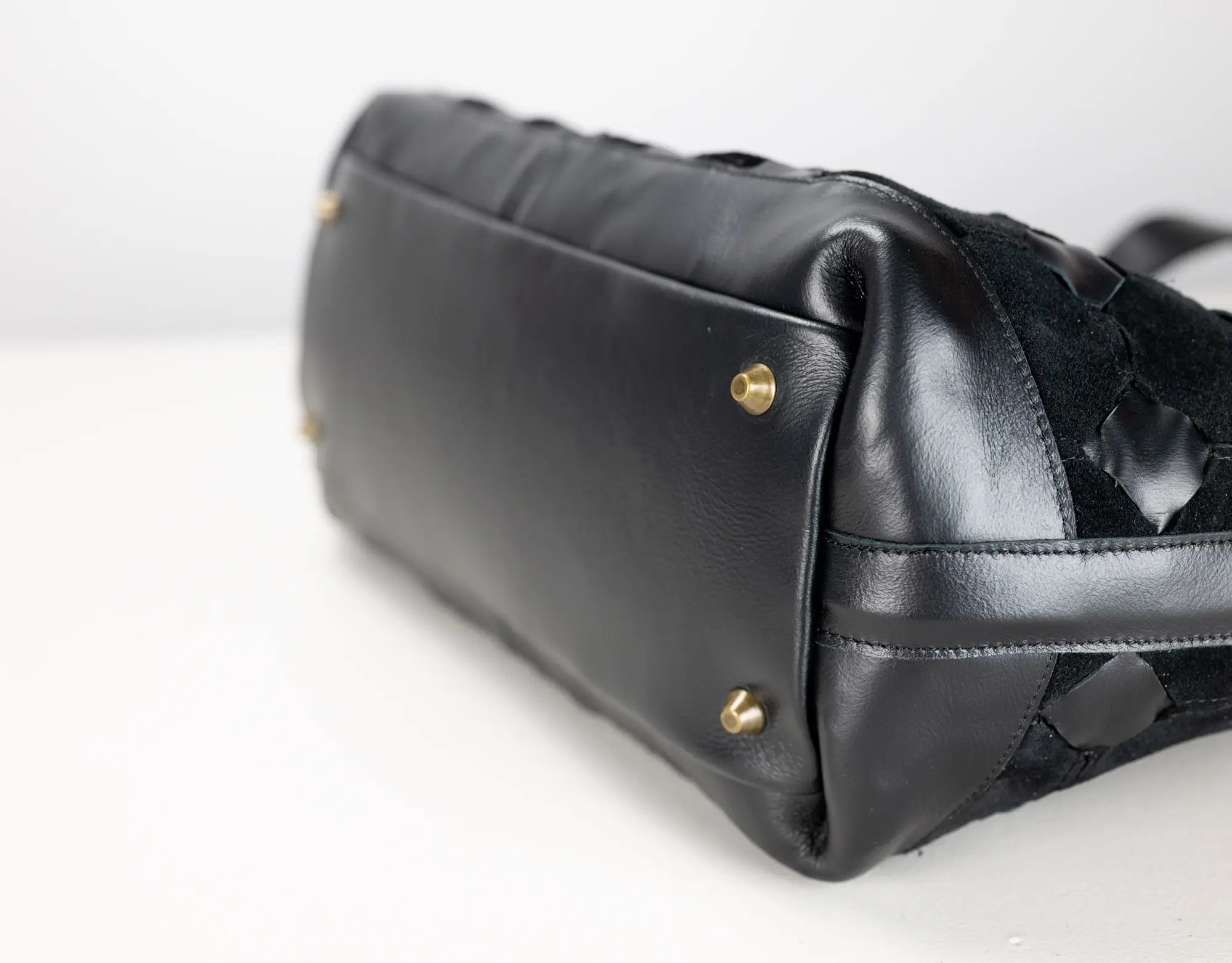 Close-up of a black leather handbag showing the bottom corner with gold-tone studs and a textured woven black strap detail on a white background.