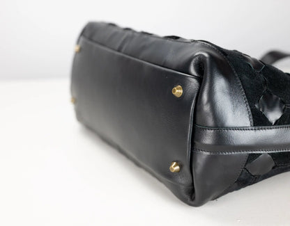 Close-up of a black leather handbag showing the bottom corner with gold-tone studs and a textured woven black strap detail on a white background.