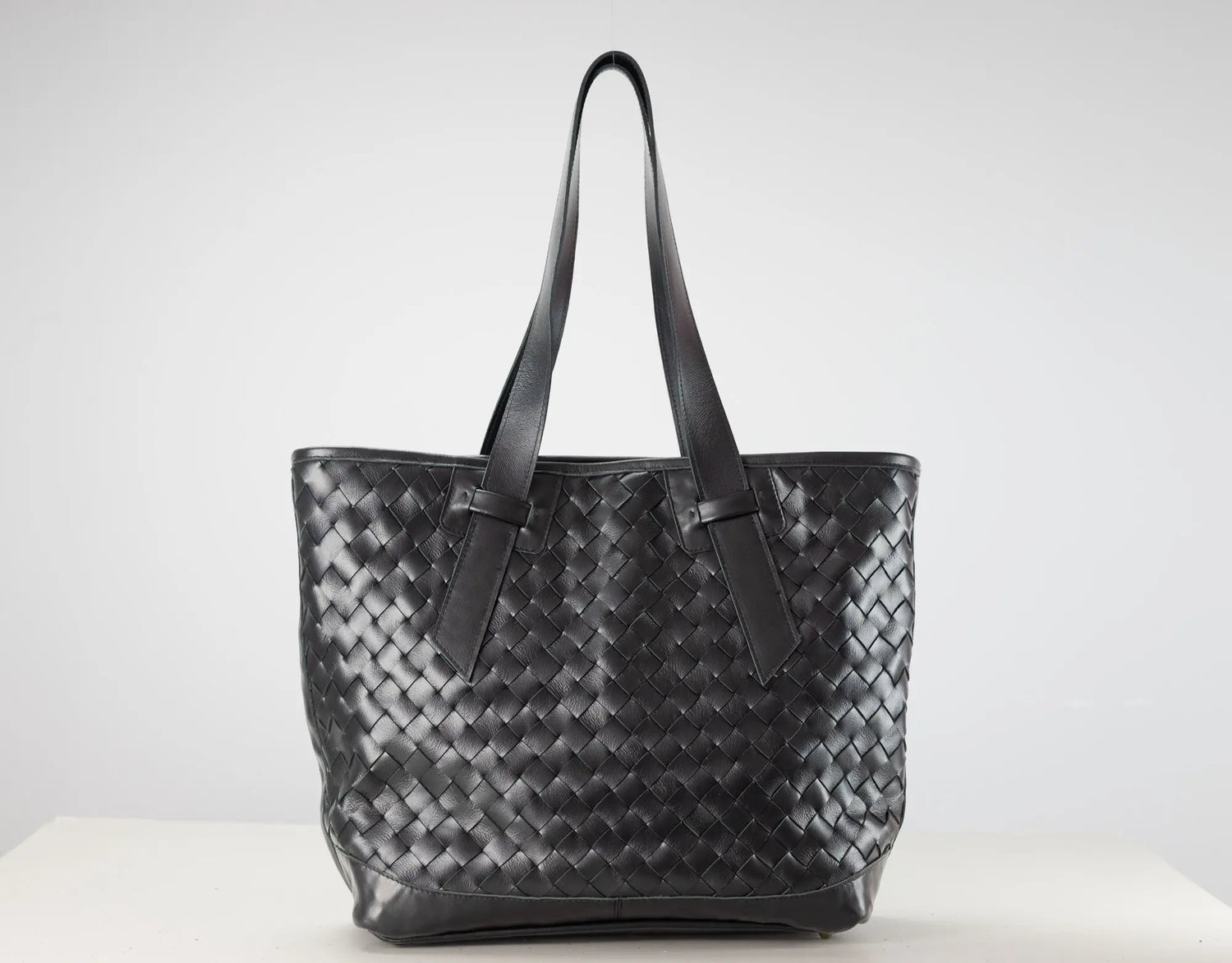 A black woven leather tote bag with two shoulder straps, displayed on a white surface against a light gray background.