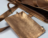 A close-up of a metallic bronze pebbled leather accessory set, featuring a small pouch, a strap, and a larger bag interior with a zipper, laid out on a light background.