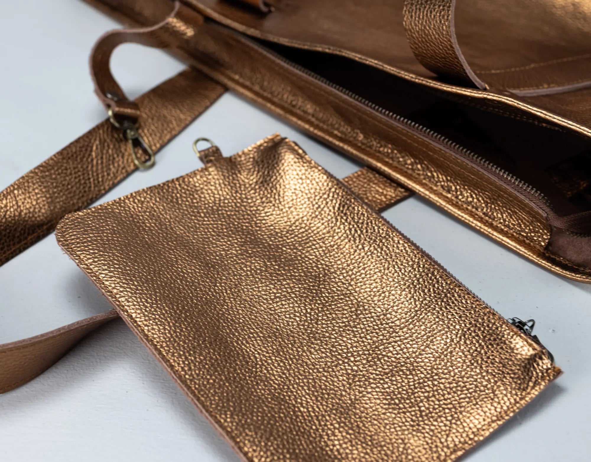 A close-up of a metallic bronze pebbled leather accessory set, featuring a small pouch, a strap, and a larger bag interior with a zipper, laid out on a light background.