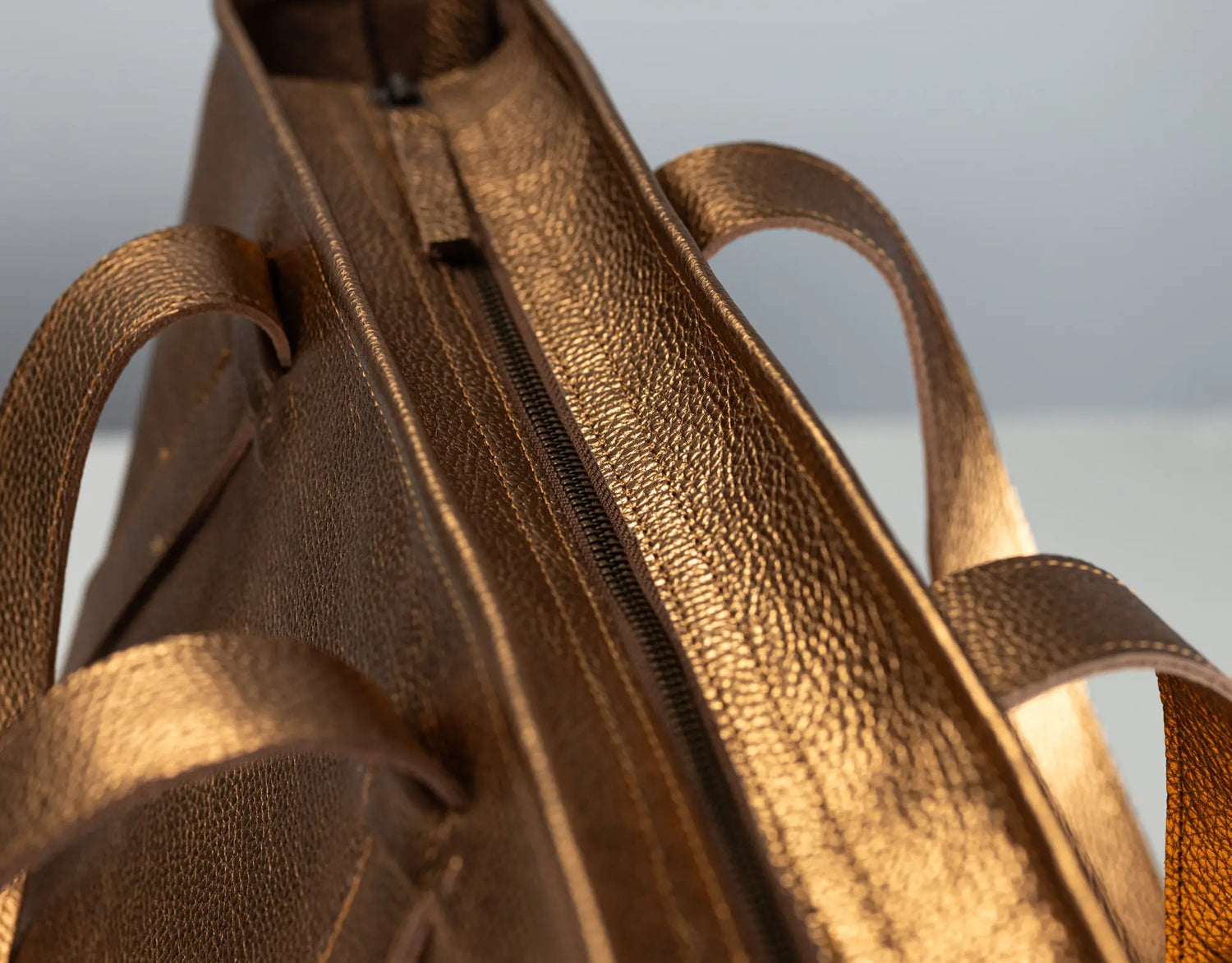 Close-up of a metallic bronze leather bag showing the pebbled texture, zipper, and part of the handle against a neutral light gray background.