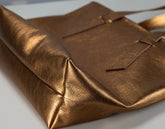 A close-up of a metallic bronze leather tote bag with a pebbled texture, showing the corner fold and strap details against a light gray background.