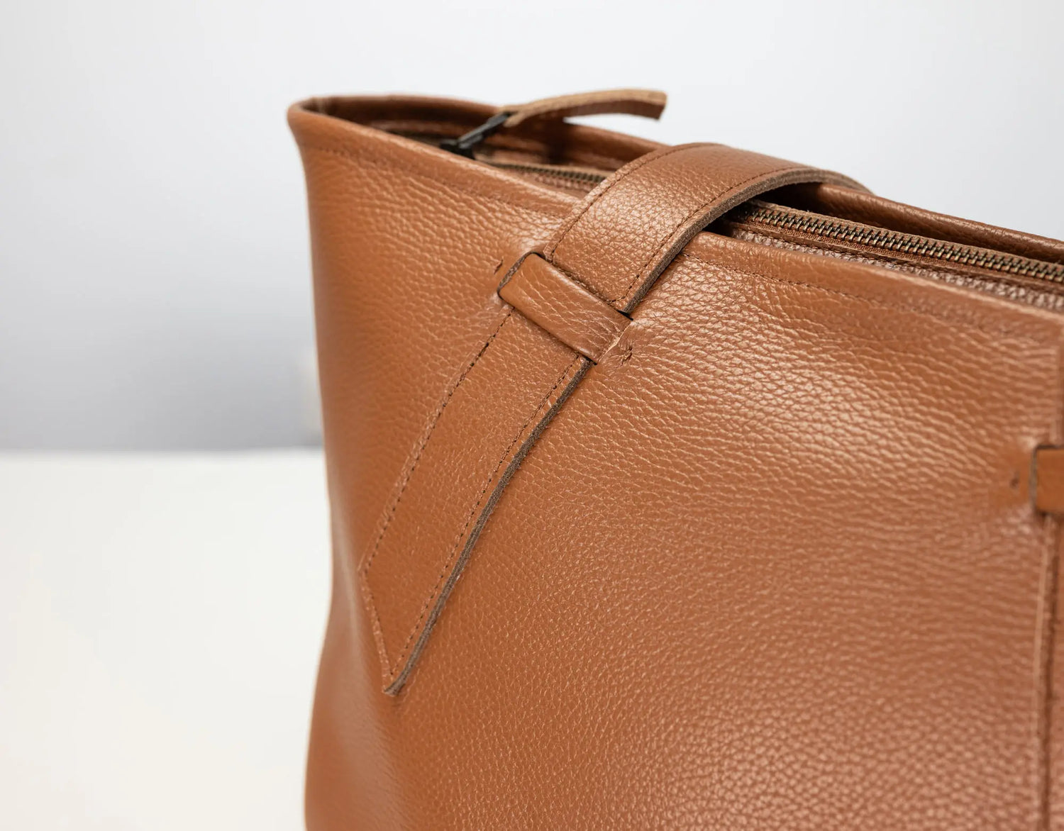 Close-up of a tan pebbled leather tote bag showing the side zipper, strap detail, and textured leather surface against a light gray background.