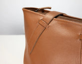Close-up of a tan pebbled leather tote bag showing the side zipper, strap detail, and textured leather surface against a light gray background.