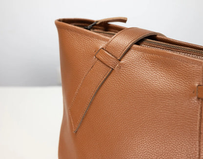 Close-up of a tan pebbled leather tote bag showing the side zipper, strap detail, and textured leather surface against a light gray background.