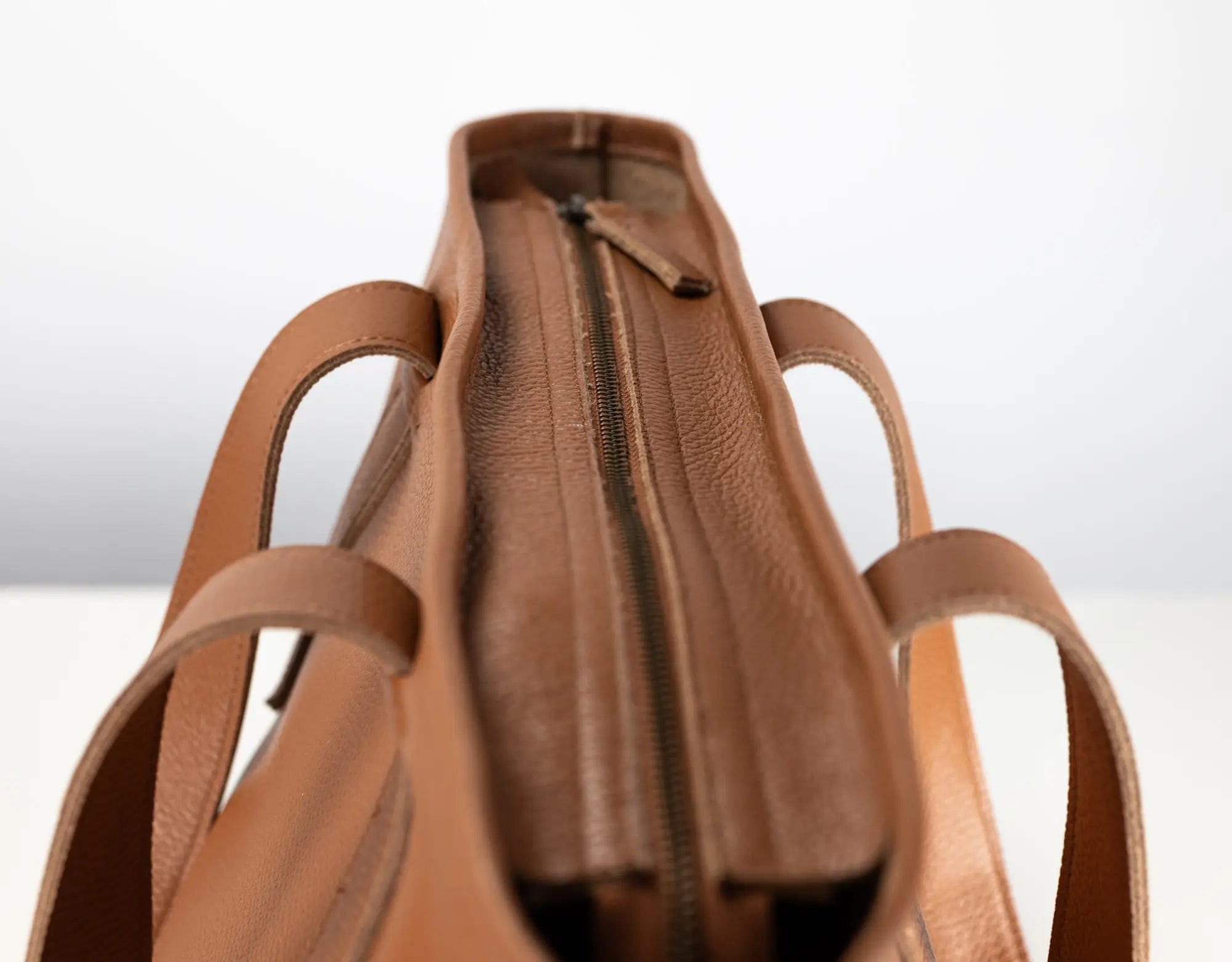 Close-up of a tan leather tote bag showing the interior zipper compartment and side handles against a light gray background.