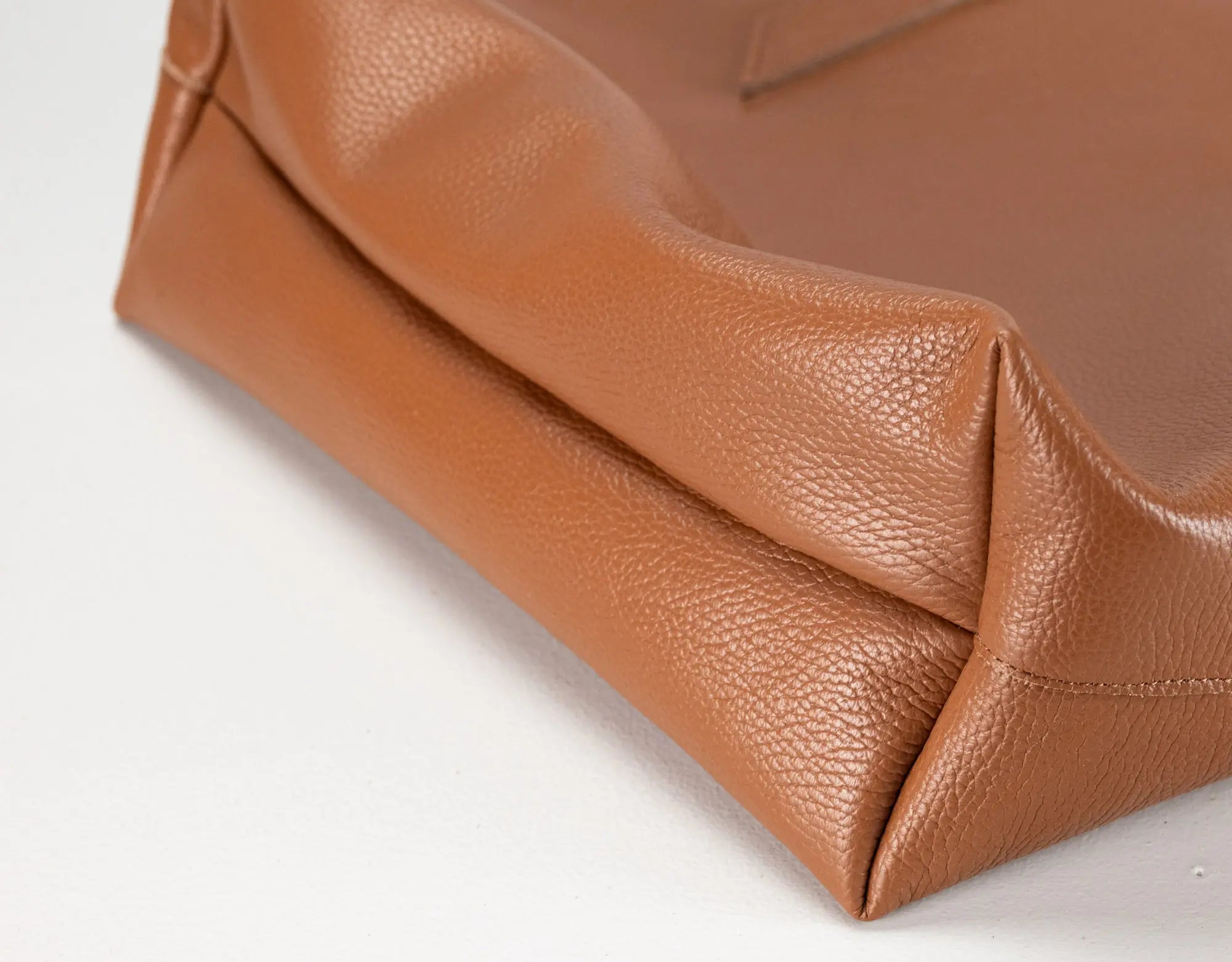 Close-up of a tan pebbled leather bag corner showing the texture and fold details on a white background.