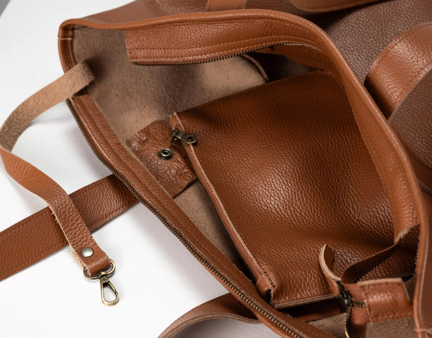 Close-up of a brown pebbled leather tote bag showing the interior compartments, zipper pockets, and strap attachments on a white background.