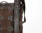 Close-up of a dark brown woven leather and suede bucket bag showing the strap attachment with brass rivets on a white background.