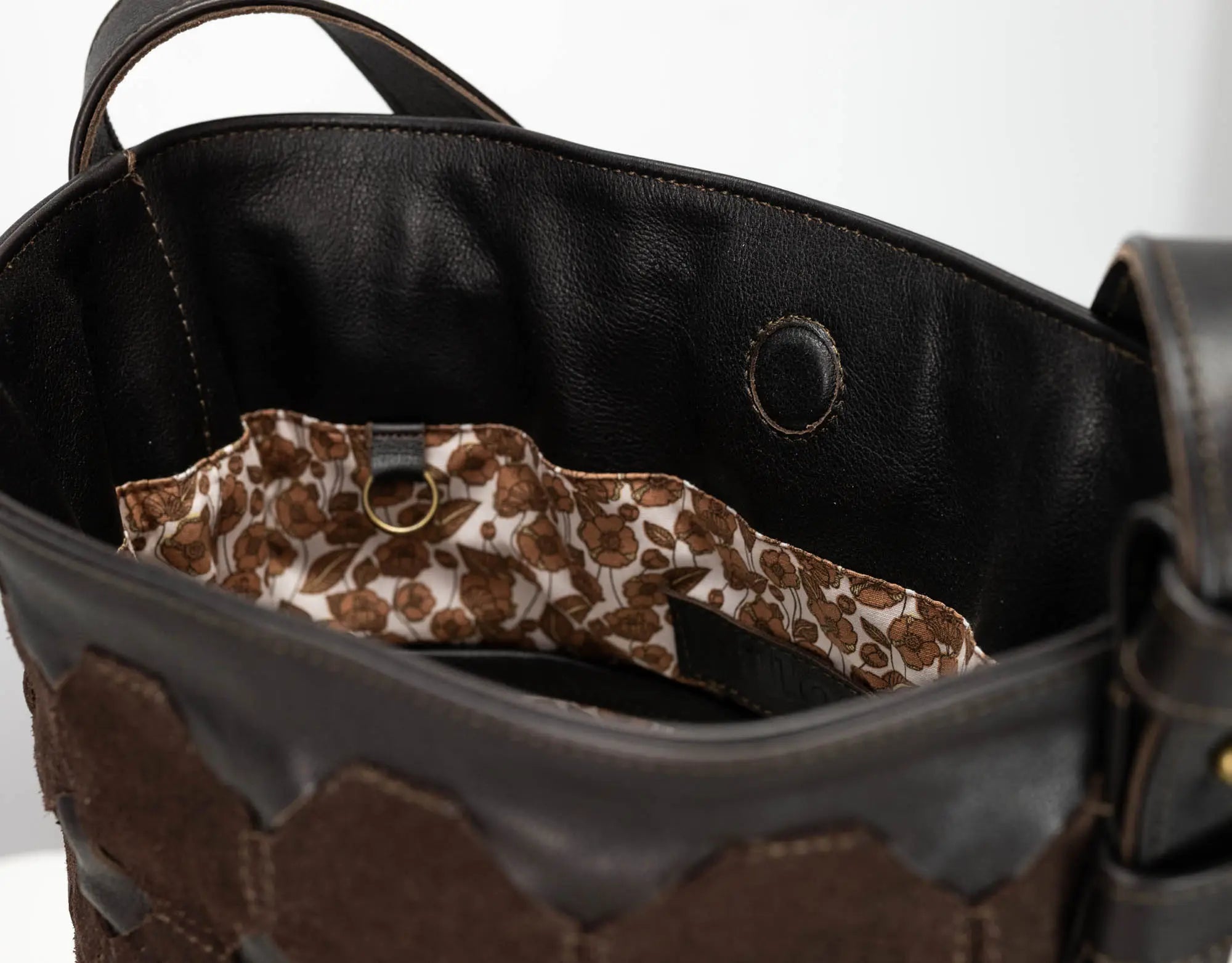 Close-up of the interior of a dark brown leather handbag, showing a black leather lining and a patterned brown and beige fabric pocket with a brass ring.