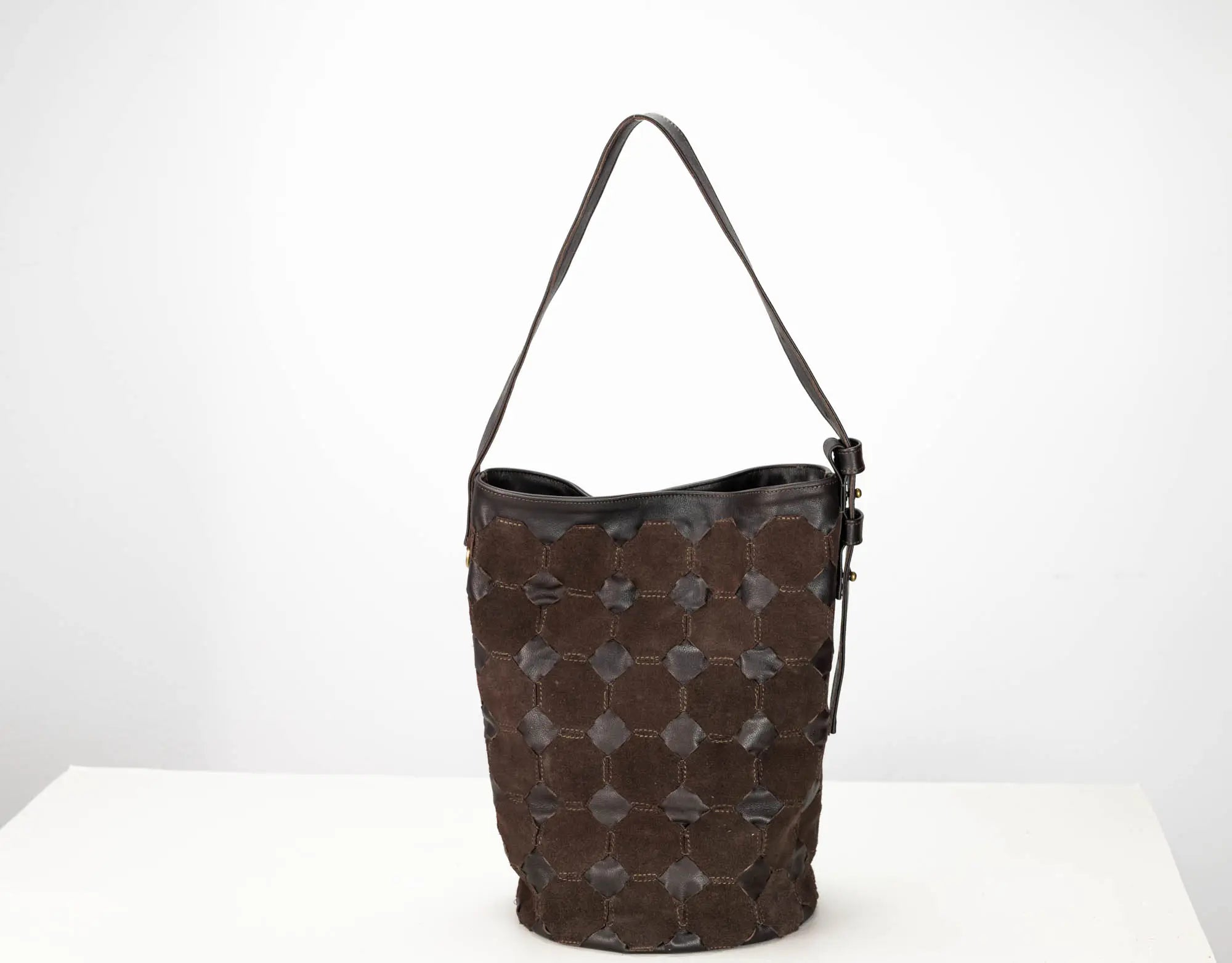 A dark brown leather bucket bag with a distinctive honeycomb pattern of stitched circular patches on the front, a black leather trim, and a single shoulder strap, set against a plain white background.