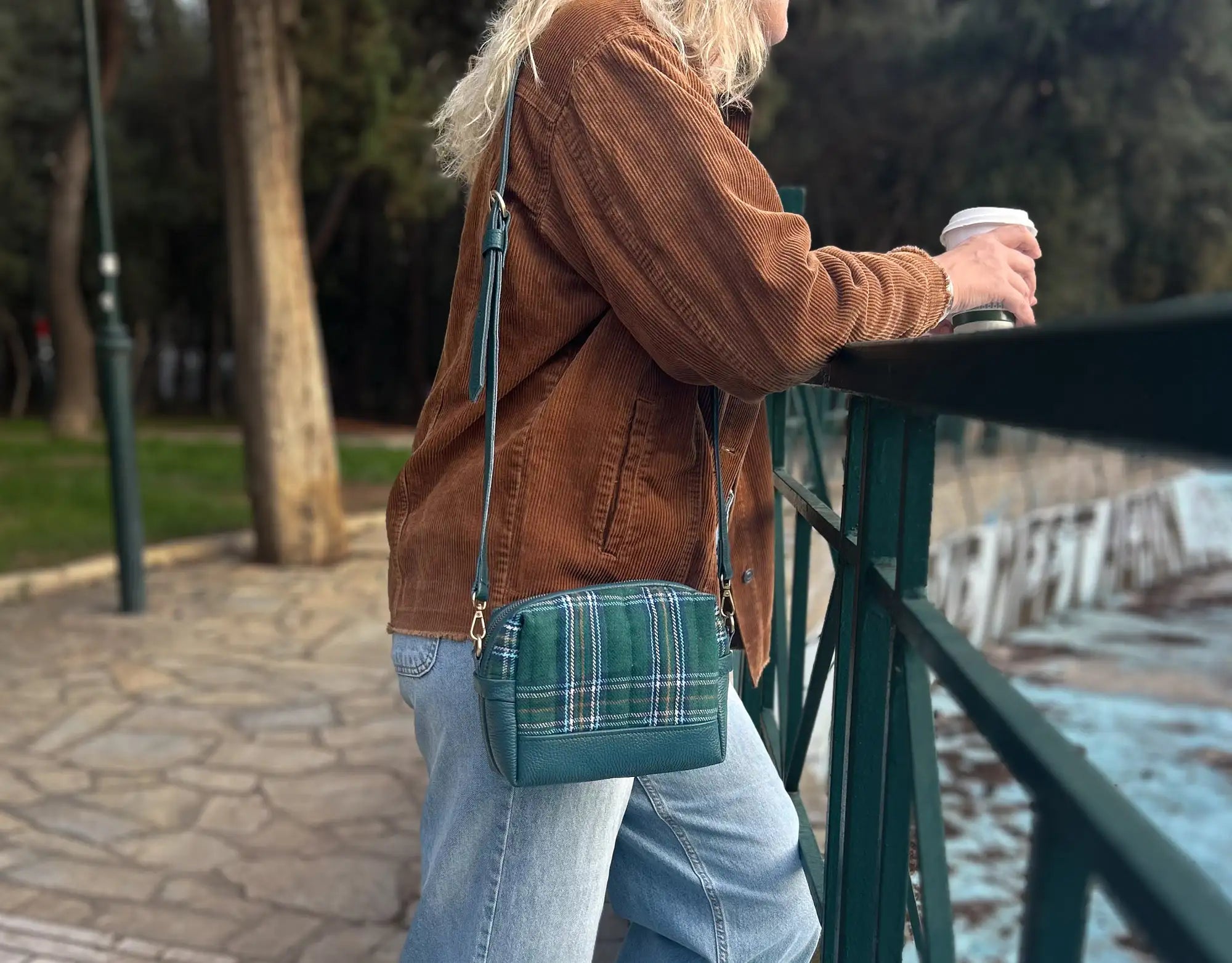 Person wearing a brown jacket and plaid bag, holding a coffee cup, standing on a bridge.